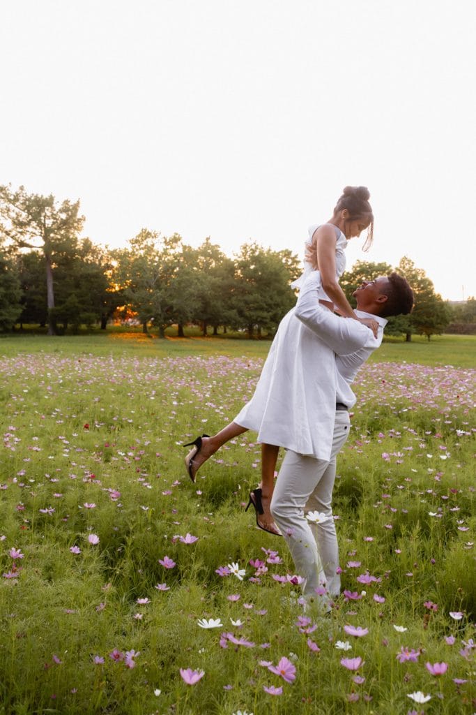 A black man lifts a black woman in the air with love in a field of pink and white flowers.