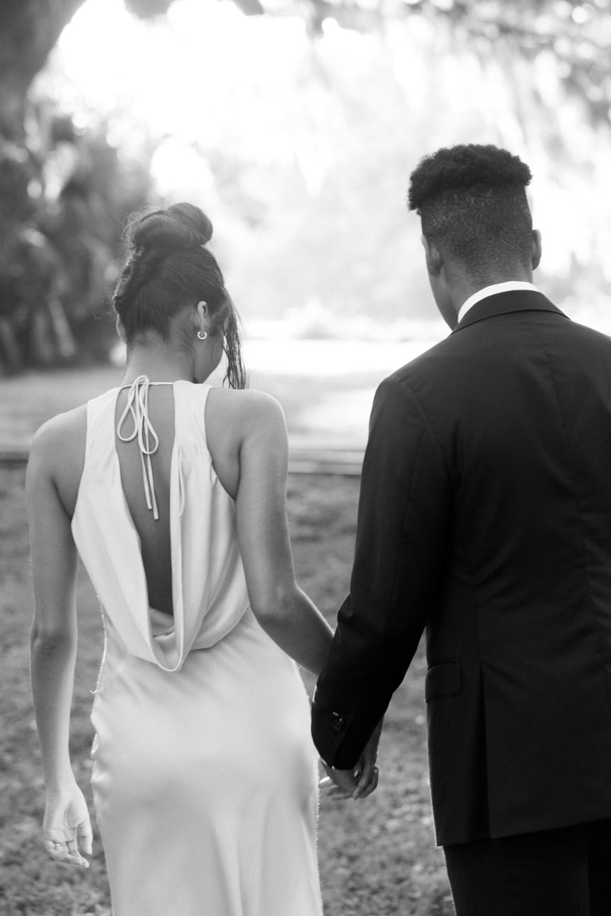 An engaged couple walks away hand in hand. The woman is wearing a backless white dress and the man is wearing a dark suit. The photo is in black and white.