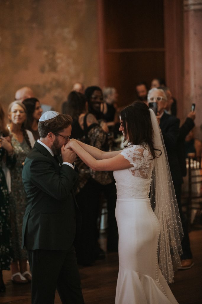 A groom in a white kippah and dark green suit kisses the brides hands during their first dance on their wedding day. She is wearing a white dress and long veil.
