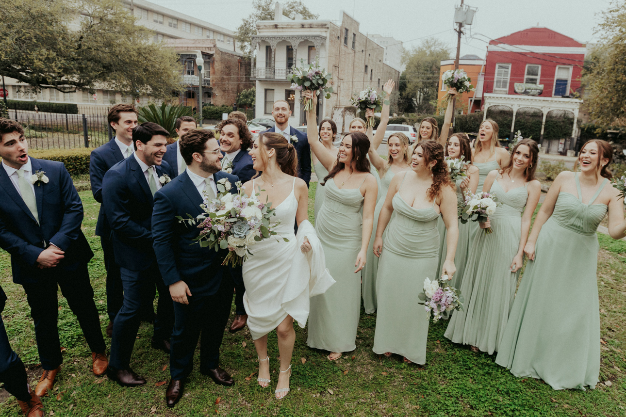 A group of bridesmaids and groomsmen and a bride and groom walk and cheer in a park in front of an old New Orleans home on their wedding day.