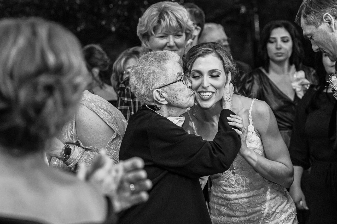 A bride's grandmother gives her a kiss on the cheek as the bride smiles at her wedding. The photo is in black and white.