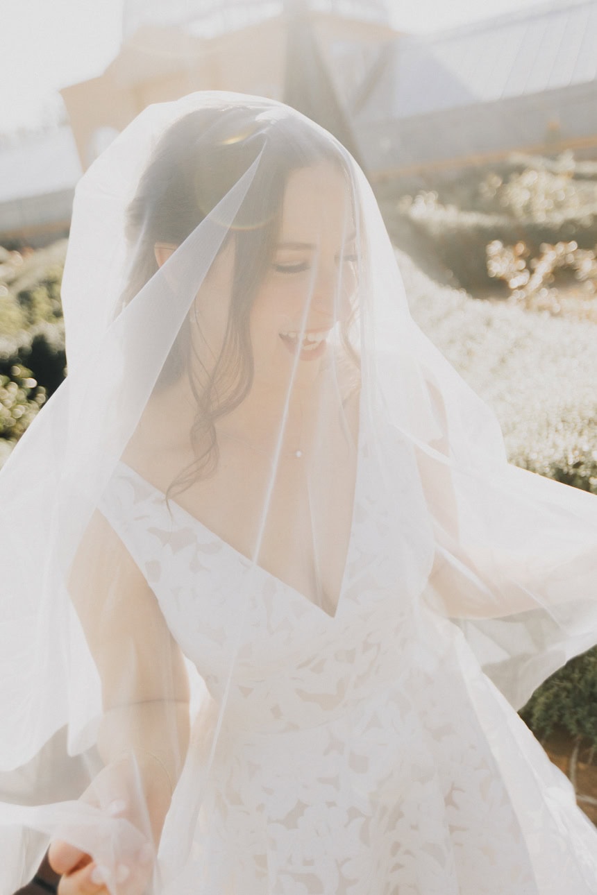 A very happy bride standing in the sunlight with her veil over her face. 