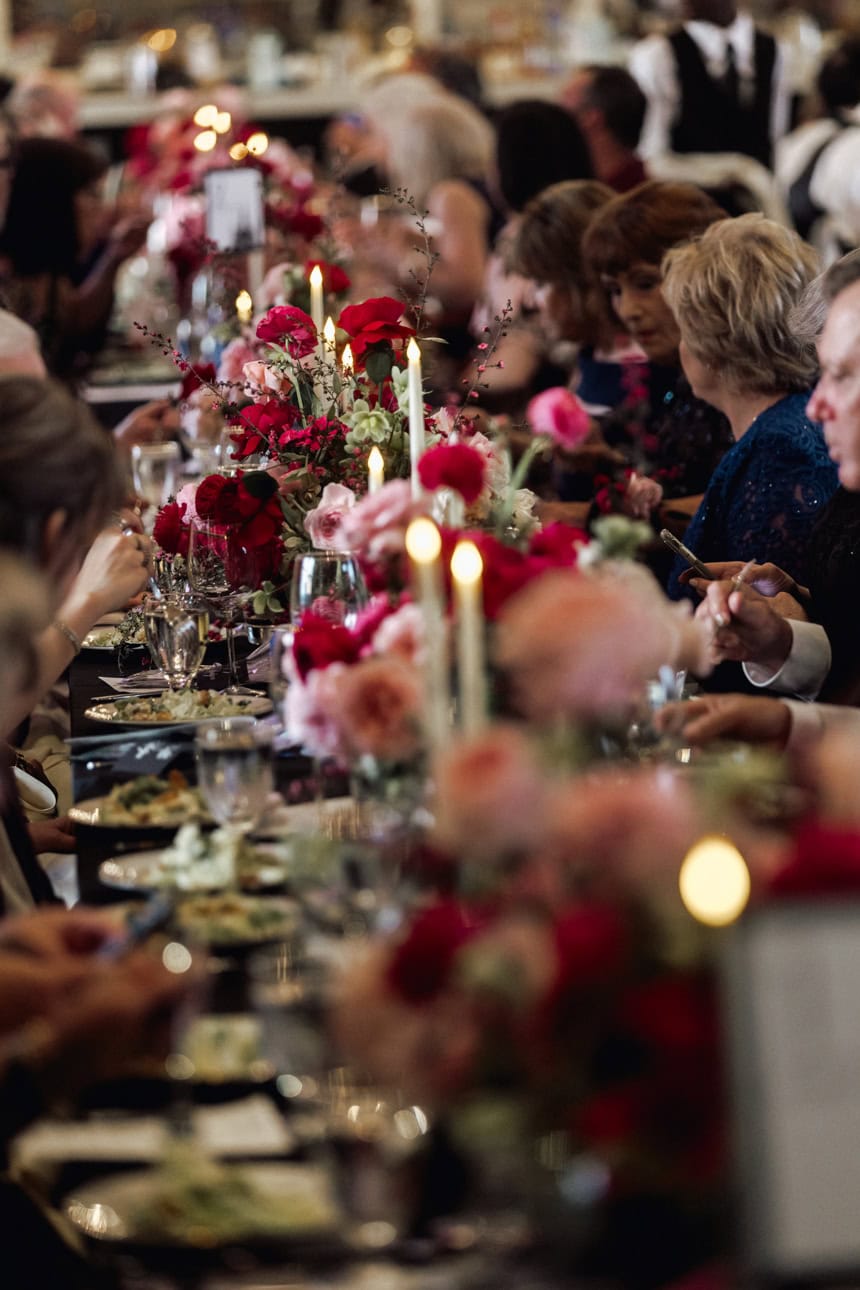Dining table at a wedding with an abundance of red and pink flowers and candlelight. Guests seated at the table enjoying dinner.