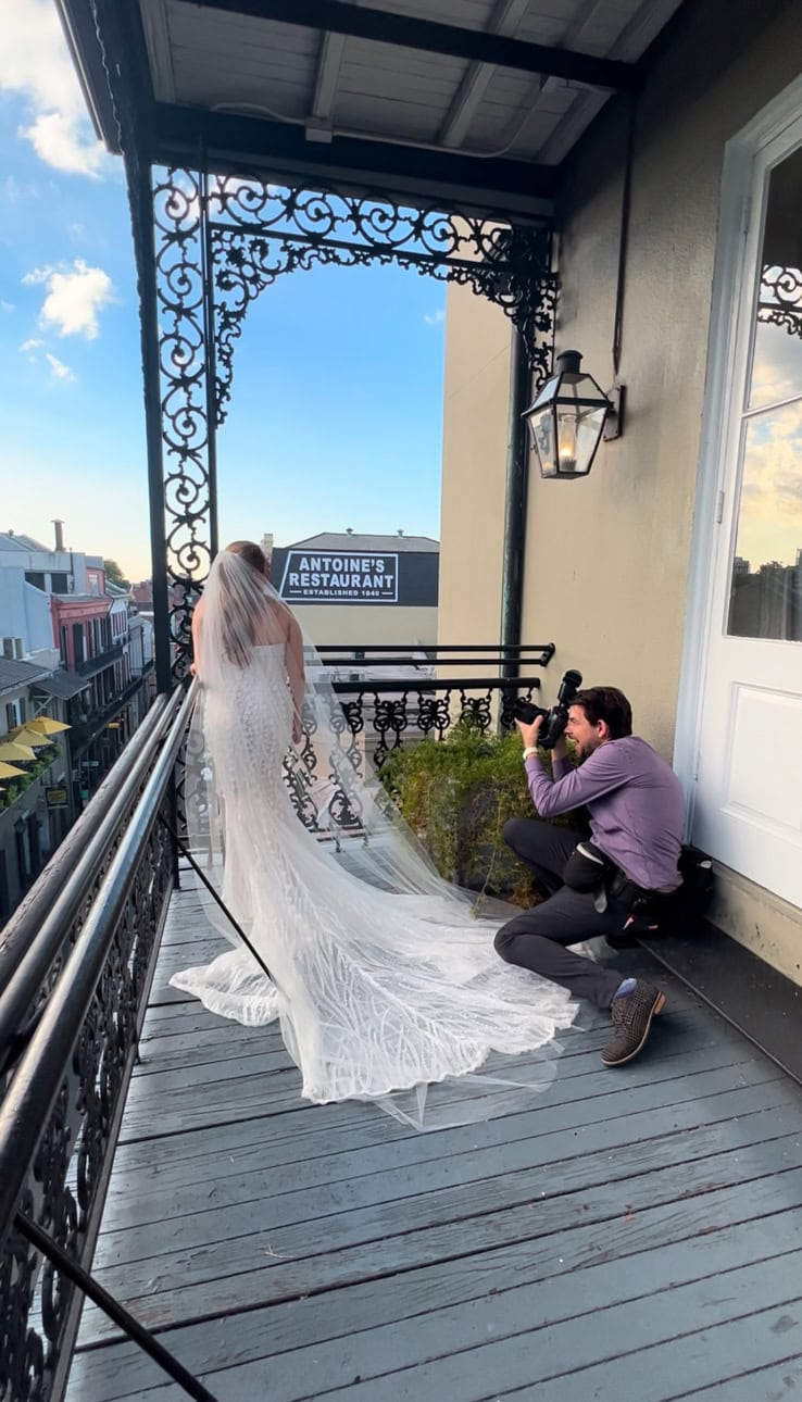 Photographer Matthew Foster kneeling down taking a photo of a bride in a white gown with a long veil on the balcony of the Omni Royal Orleans in New Orleans.