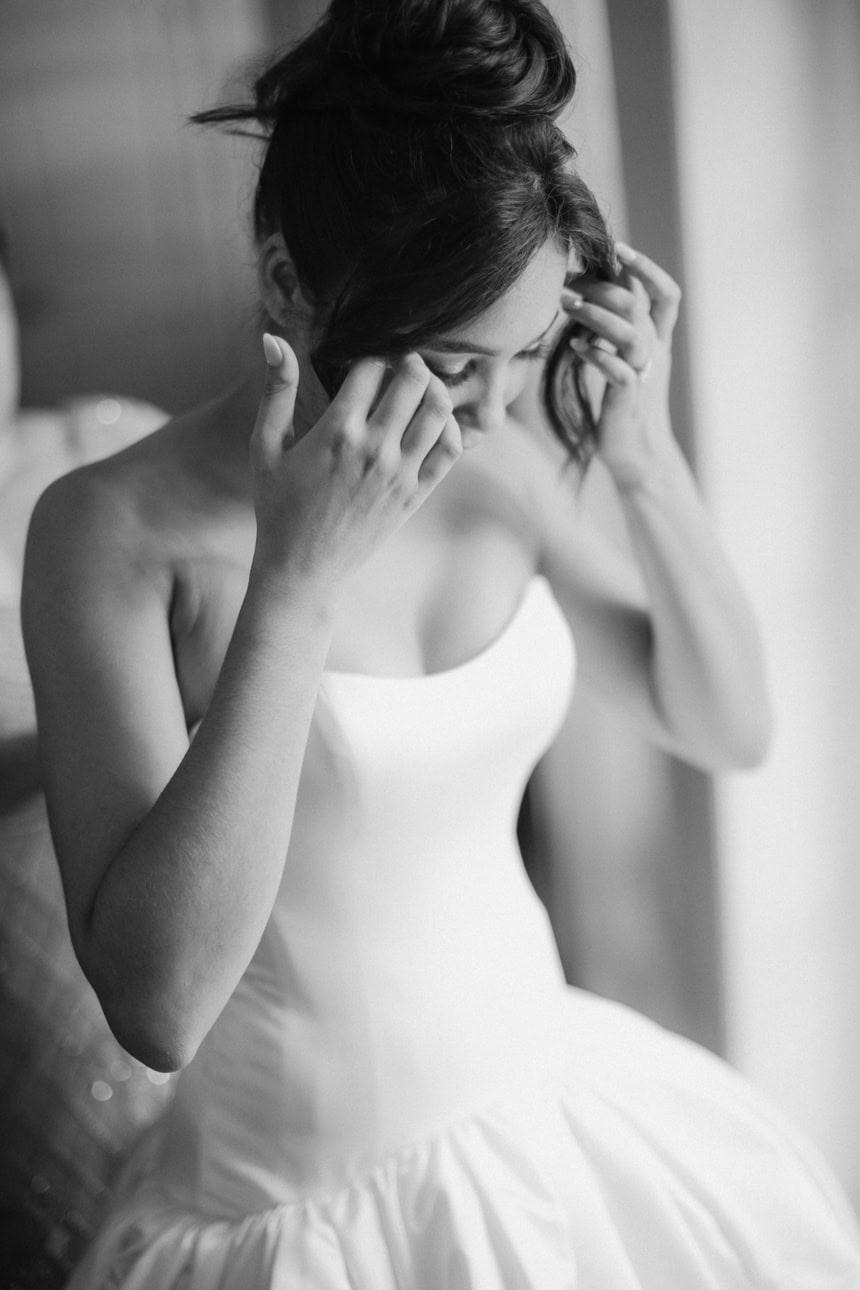 A bride adjusts her hair while looking down at her dress. The photo is in black and white.