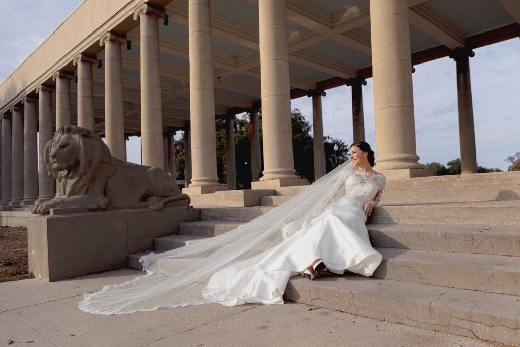 Bride in white wedding dress with long veil sitting on stone steps under a classical building with lion sculpture background.