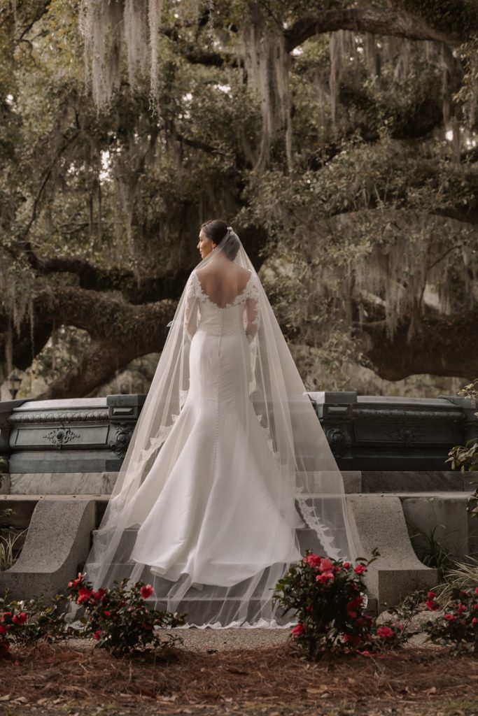 Bride in white wedding gown with train and veil stands on stone bench.