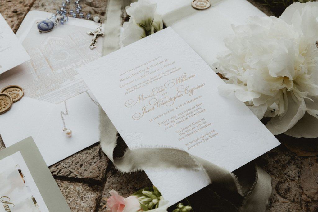 Charming wedding invitation details with floral decorations, pearls, and religious icons on a rustic stone surface.