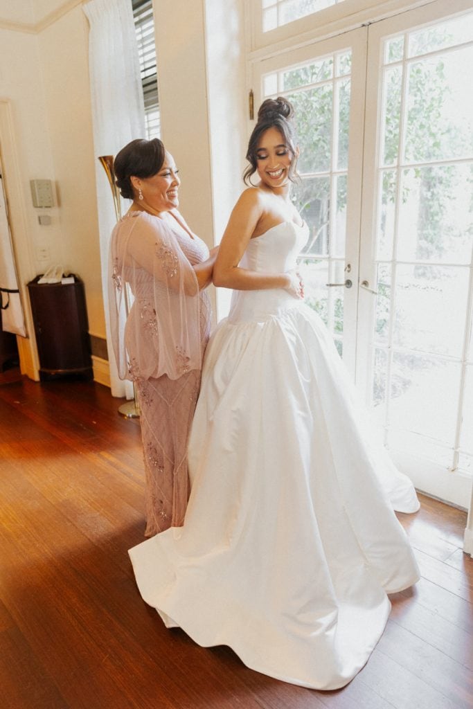Beautiful bride smiling in a white wedding gown with her mother preparing her for the wedding.