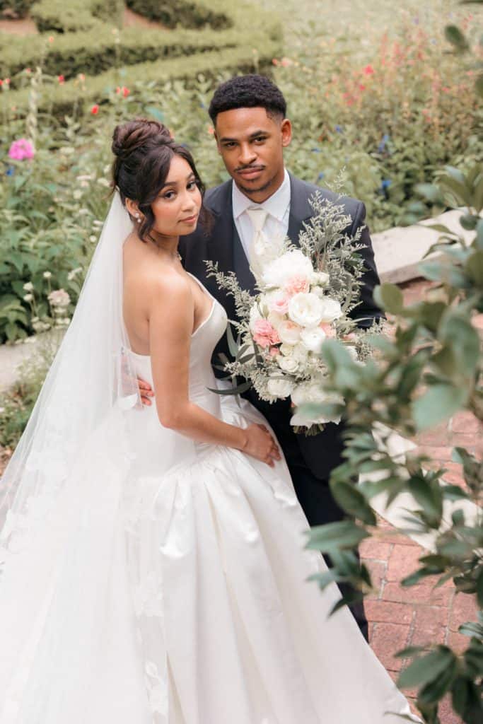 Elegant bride and groom posing with a soft-focus floral background, wedding attire, and bouquet.
