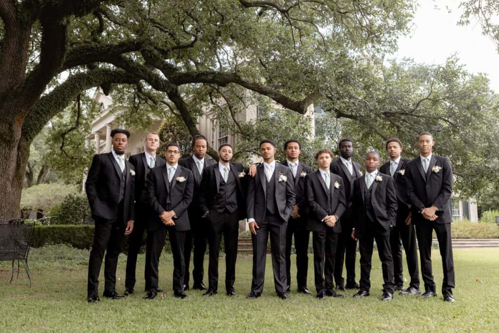 Group of groomsmen in black suits and ties standing under a large tree in a garden setting, celebrating a wedding in New Orleans.