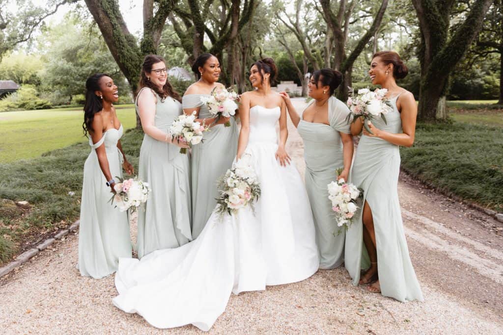 Bright, joyful wedding bridesmaids and bride with flowers in a lush park setting.
