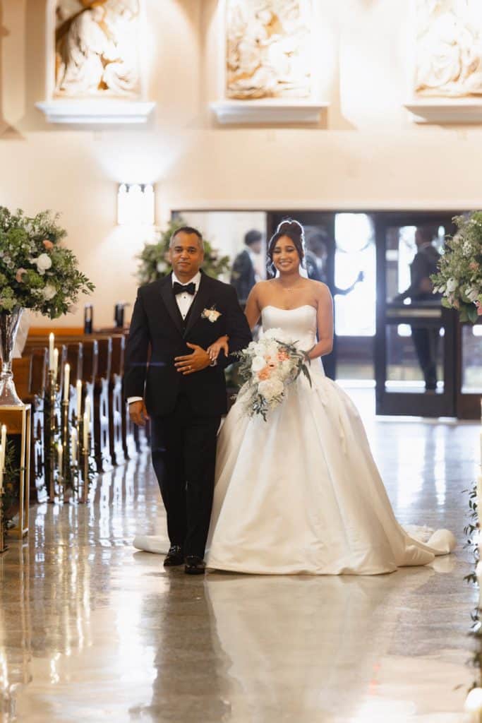 Bride in white wedding gown holding bouquet, walking with father in a formal black tuxedo inside church.