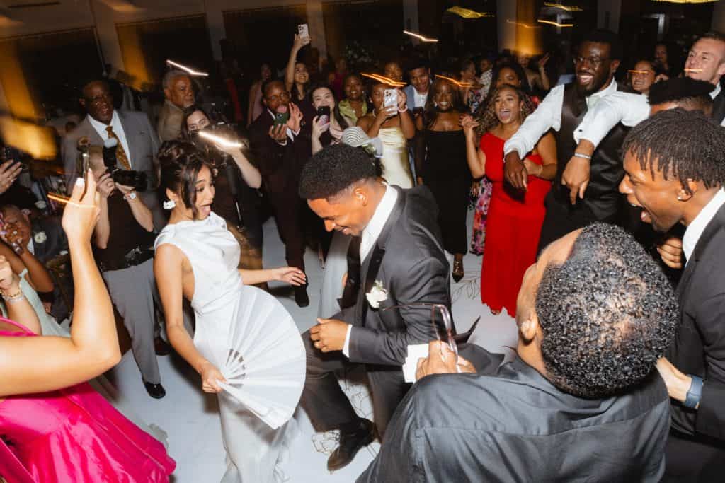 Joyful couples dancing at a lively wedding reception with guests capturing moments on smartphones.