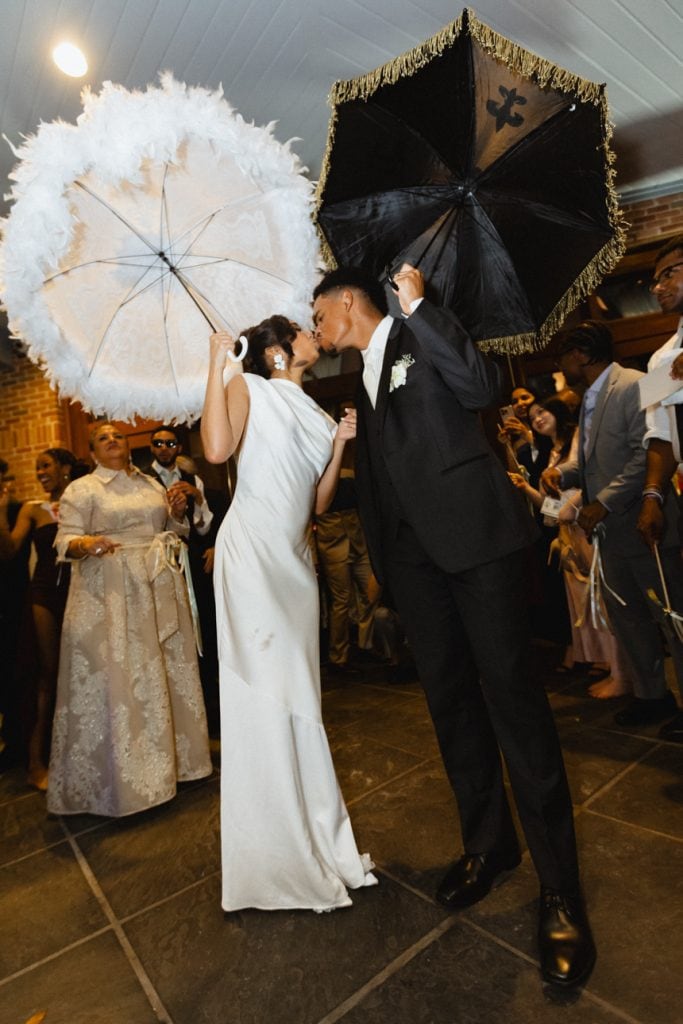 Vibrant wedding moment in New Orleans with bride and groom kissing under ornate umbrellas, surrounded by joyful guests.