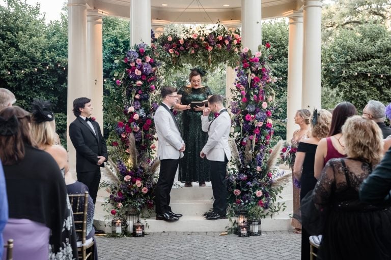Two grooms exchange vows at the gazebo at the Elms Mansion in New Orleans. One groom has been moved to tears during this emotional moment. Colorful florals frame the gazebo.