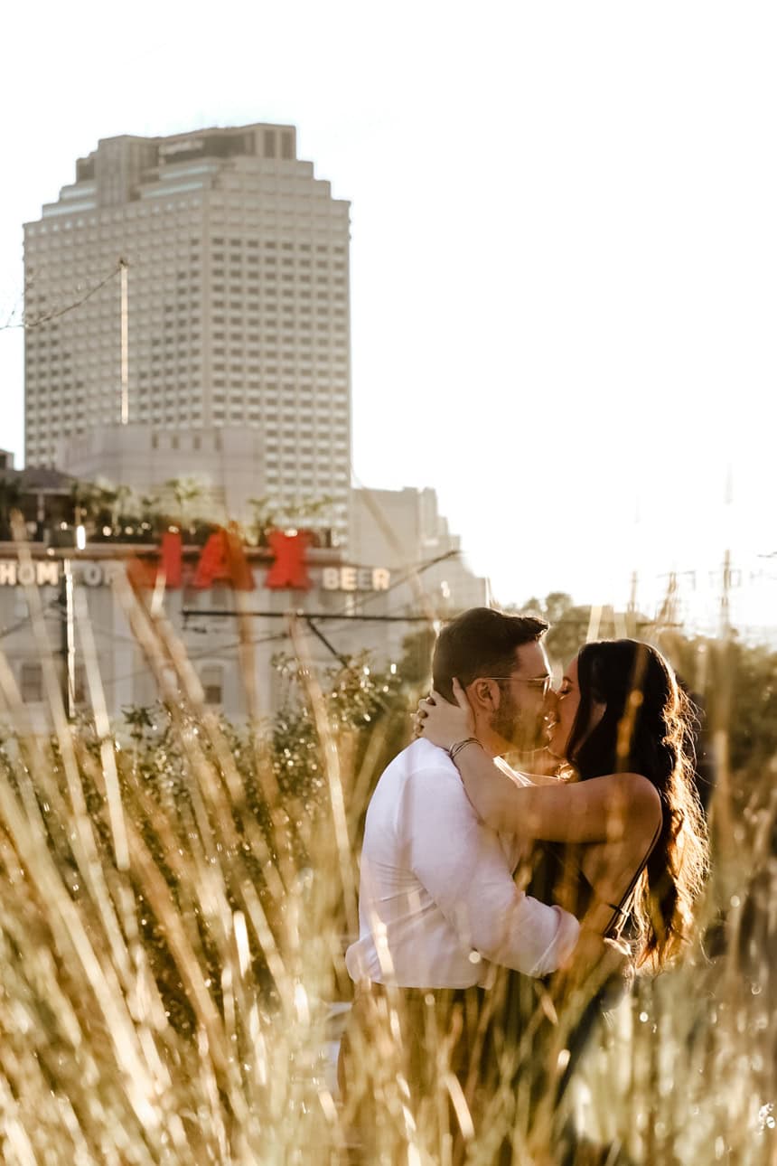 A couple embrace in the sunlight with the New Orleans skyline behind them.