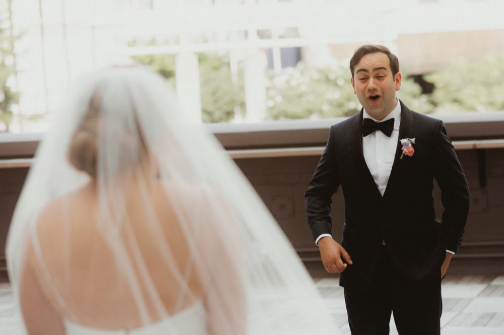 A groom in a tuxedo reacts happily to his bride during their first look.
