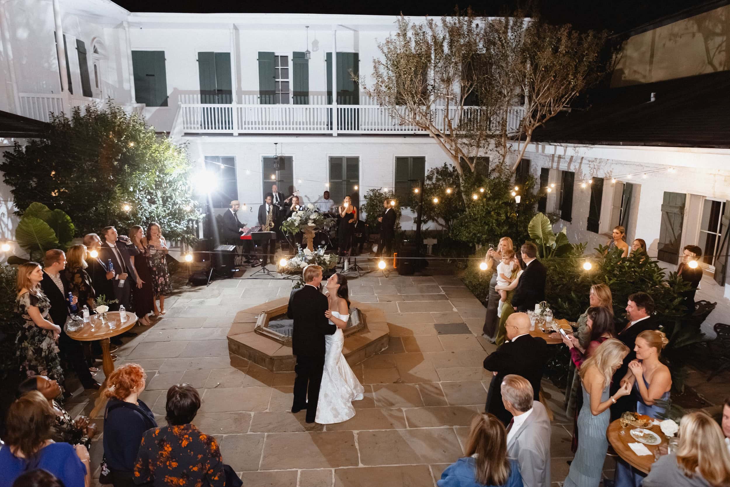 Romantic couple sharing their first dance surrounded by friends and family under string lights at the BK House.