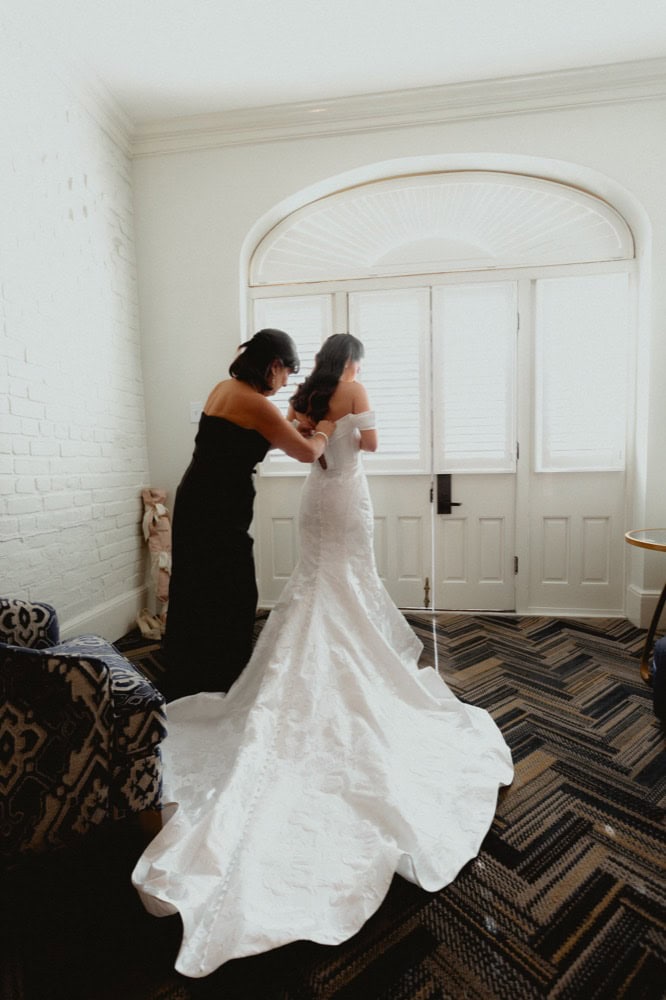 Bride in a white wedding gown with a long train, standing by a window.
