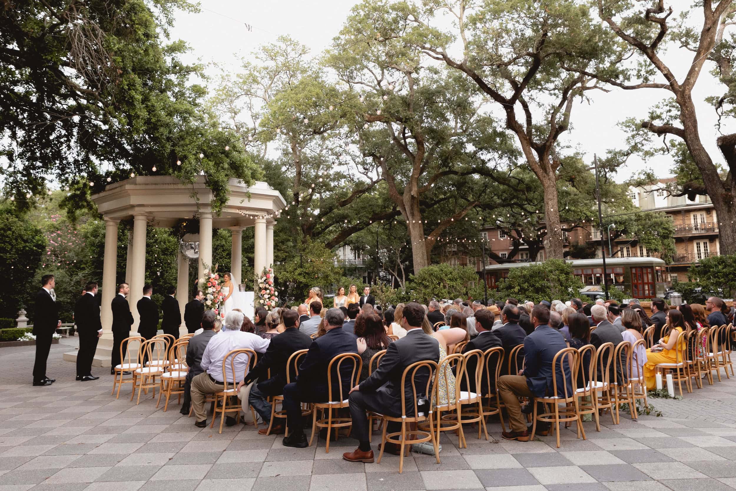 Wedding ceremony at The Elms Mansion in New Orleans with guests seated outdoors under large trees with the streetcar going by in the background.