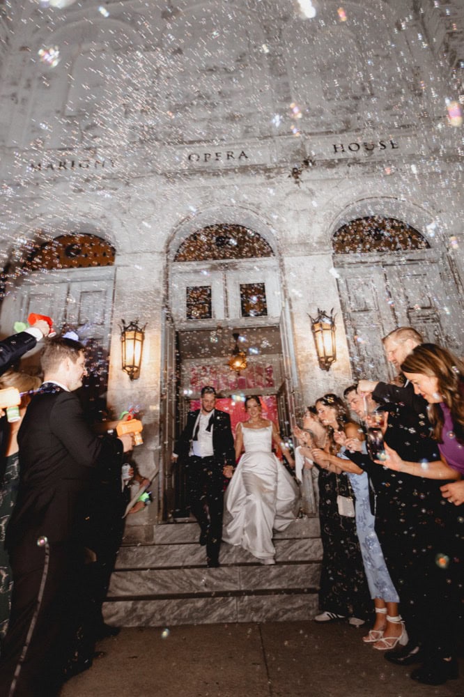 Bride and groom exit their wedding reception outside the historic Marigny Opera House in New Orleans, the air filled with bubbles.