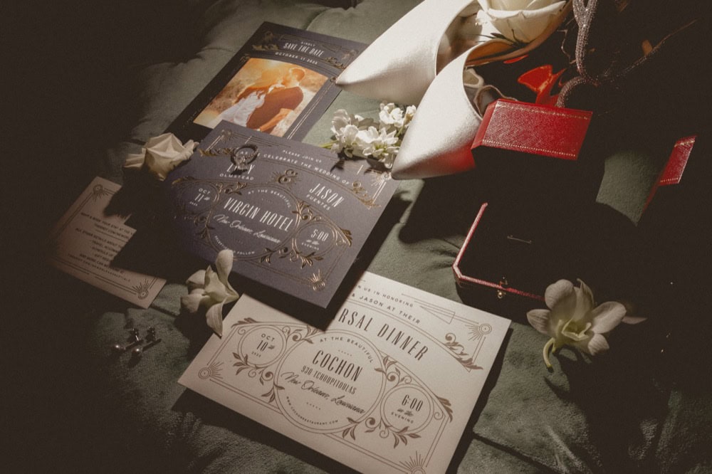 Stylish wedding invitations laid out with white high heeled shoes and a red ring box on top of a green background.