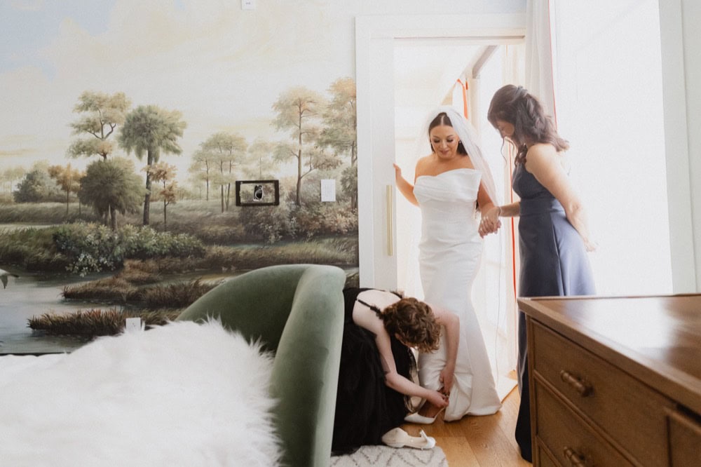 A bridesmaid helps a bride put her shoes on in a room at the Virgin Hotel in New Orleans. The bride's mother holds her hand.