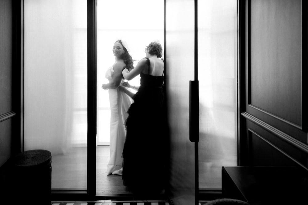 A bridesmaid assists the bride by buttoning the back of her dress as they stand in a frosted glass doorway in a room at the Virgin Hotel in New Orleans.