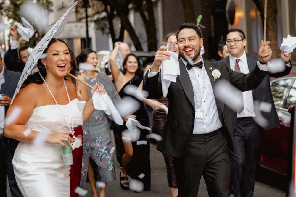 Bride and groom celebrating in New Orleans during a second line parade, confetti flies through the air.