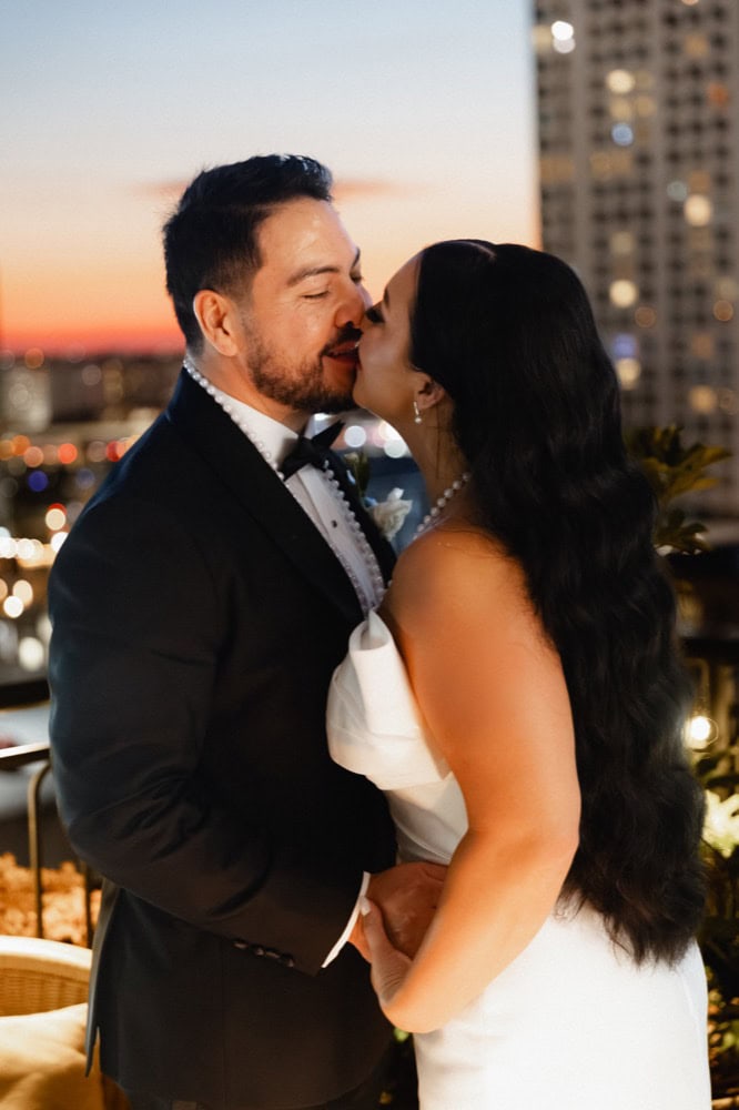 The bride and groom share a kiss on the balcony of the Virgin Hotel in New Orleans, the sunset and city lights behind them.