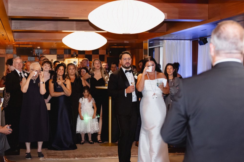 The bride wipes a tear away as she and the groom and the wedding guests listen to a wedding toast given by her father.