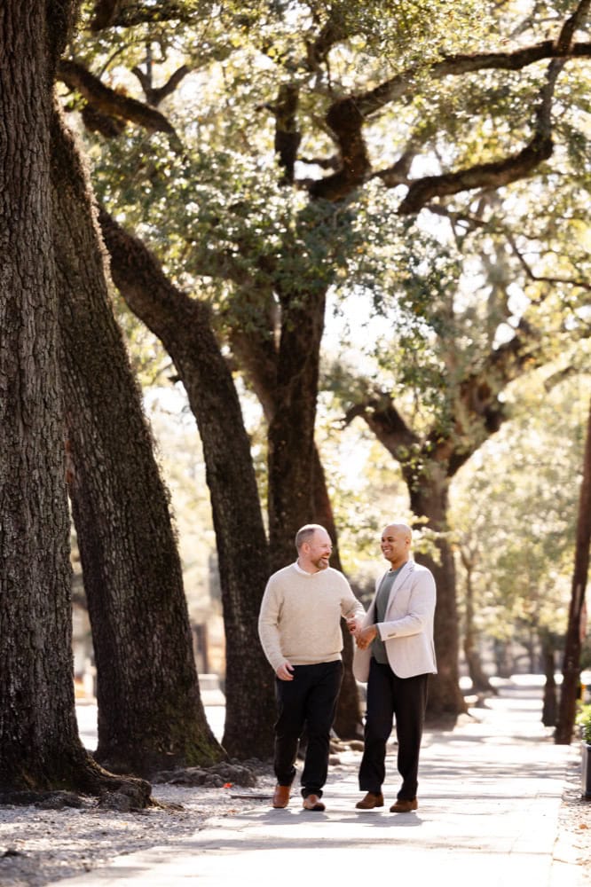Two men holding hands in a park with large trees and sunlight filtering through the leaves.
