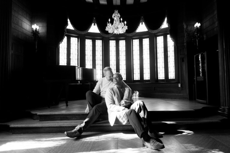 Couple sitting on stage in elegant venue, the Elms Mansion, with chandelier and large windows.