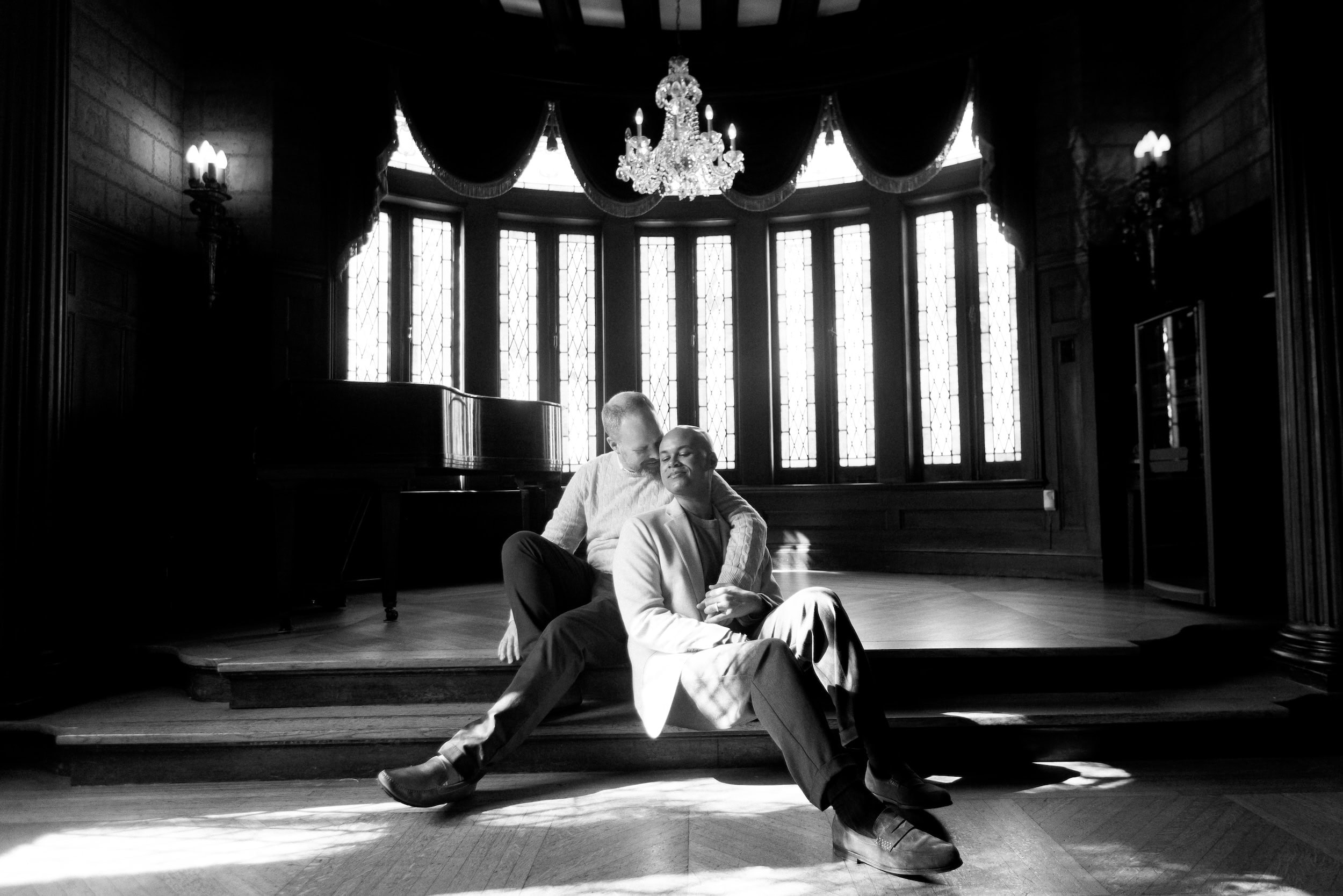 Couple sitting on stage in elegant venue, the Elms Mansion, with chandelier and large windows.