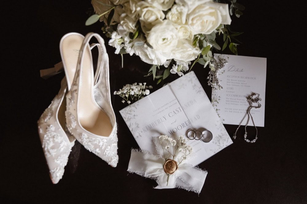 Wedding shoes and jewelry arranged with flowers and wedding invitation.