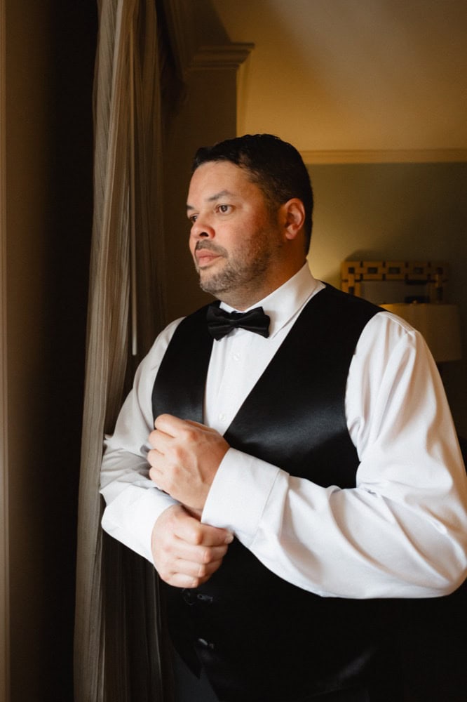 Groom in tuxedo gazing out window, preparing for his wedding in New Orleans.
