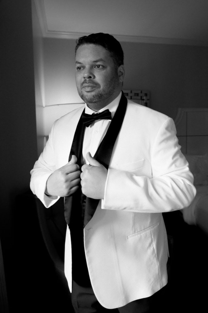 Groom in white tuxedo adjusting his bow tie before wedding ceremony.