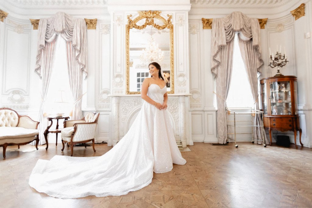 Bride in wedding gown in luxurious historic ballroom with ornate decor.