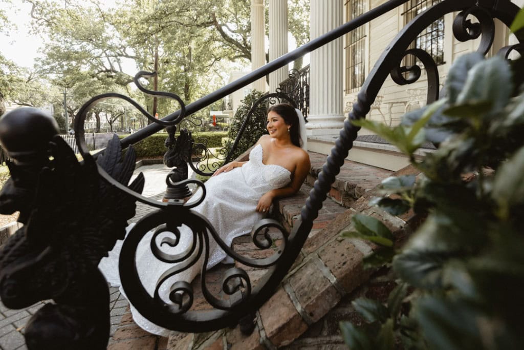 Bride in white wedding dress sitting on porch steps with ornate railing.