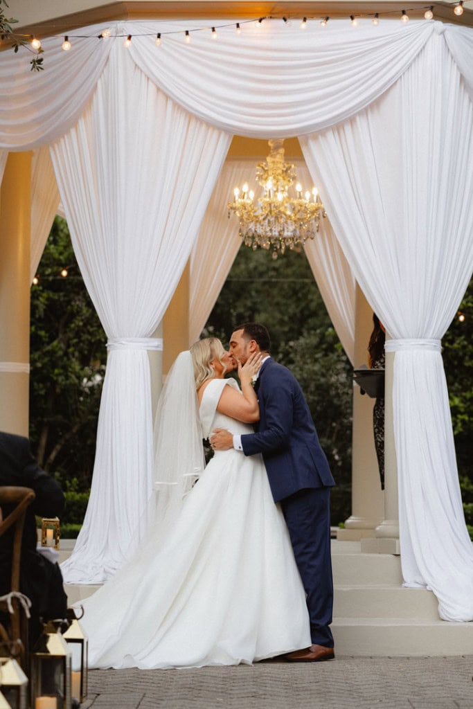 Bride and groom kissing under elegant white drapery with chandelier in New Orleans.