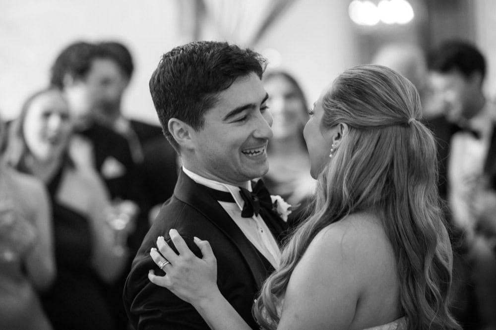Couple dancing at wedding with guests in background, black and white.
