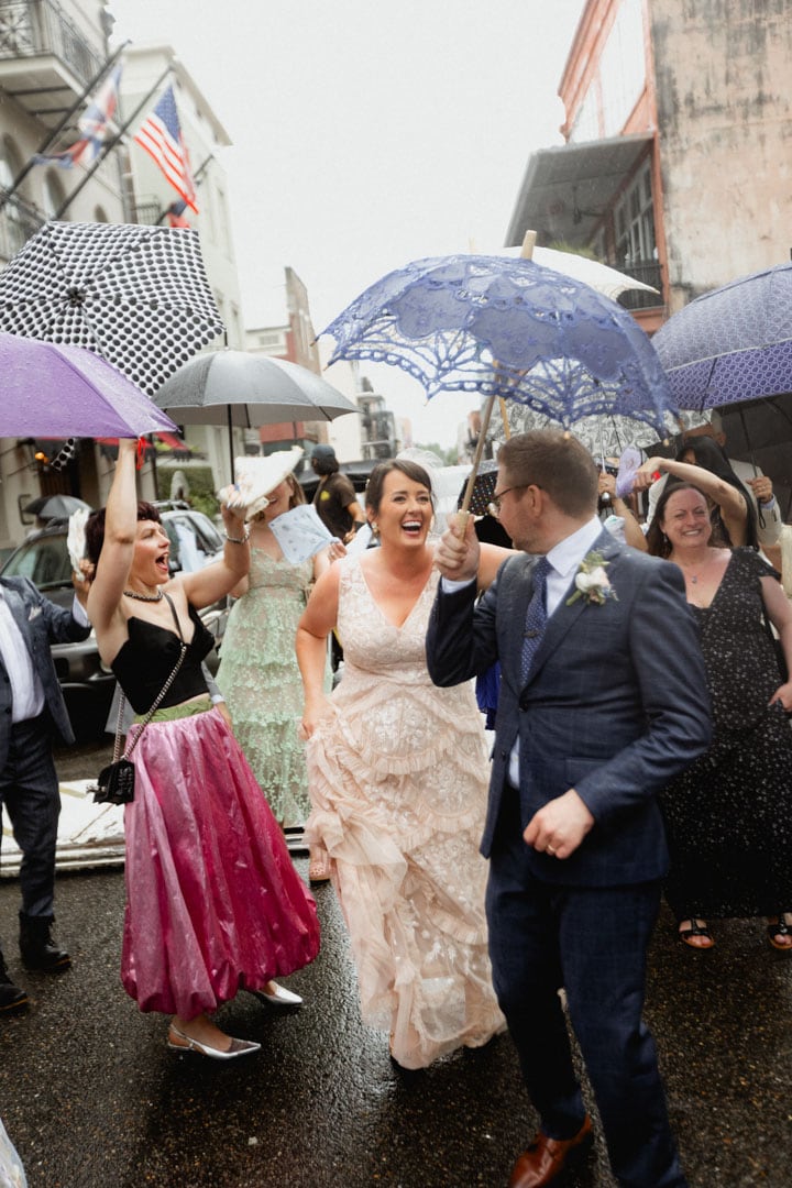 Bride and groom celebrating with guests under umbrellas in New Orleans during their rainy second line parade.