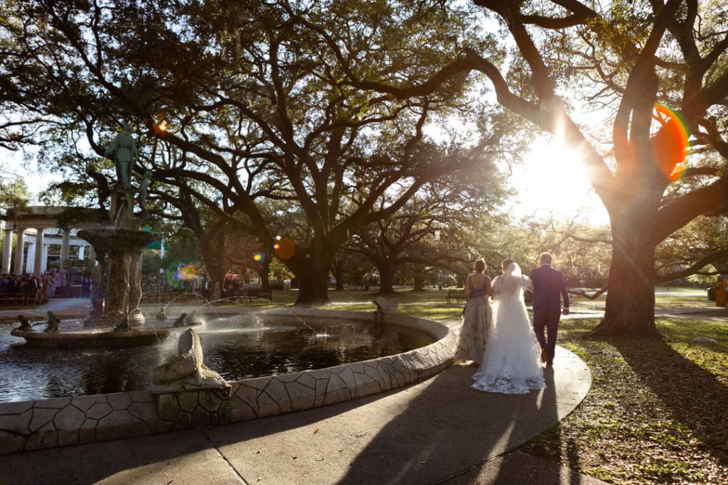 Bride with her parents walking in the Audubon Zoo with fountain and large trees at sunset.