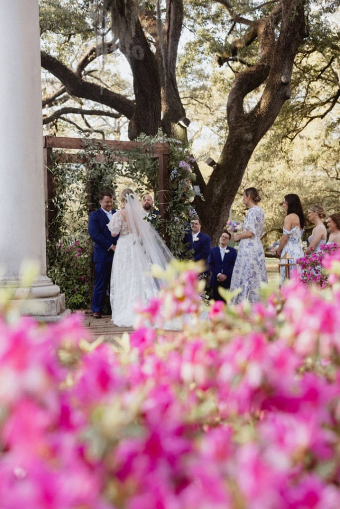 Wedding ceremony outdoors with bride, groom, and guests under large oak tree.