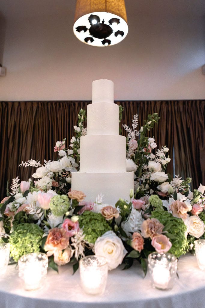 White towering wedding cake surrounded by pink and white flowers and white candles